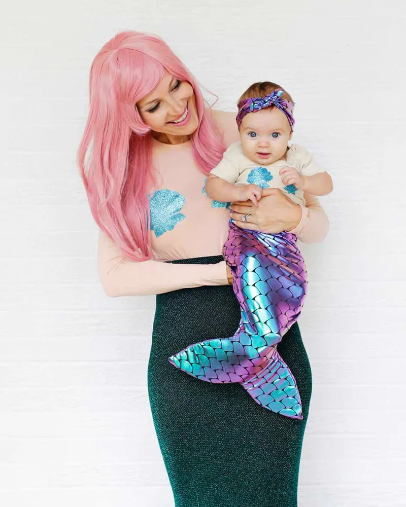 Mother with a pink wig holding her baby dressed in a shimmering mermaid tail and shell top