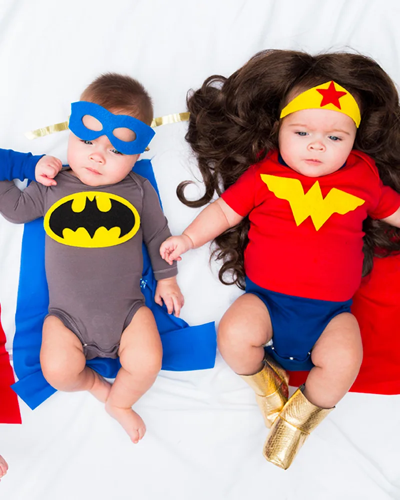 Two babies in superhero onesies with felt masks, capes and a big curly wig