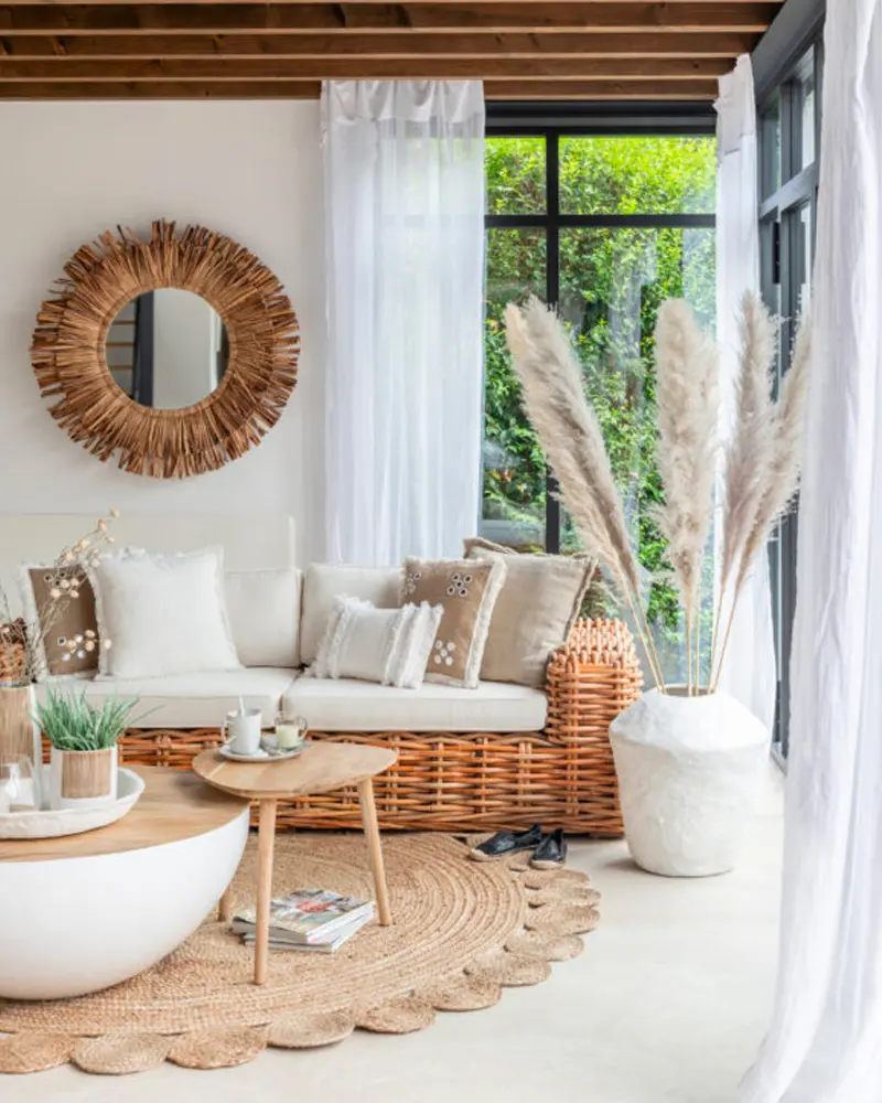 Boho living room with a rattan sofa, a sunburst mirror, pampas grass in a white vase, a round jute rug and a white drum coffee table