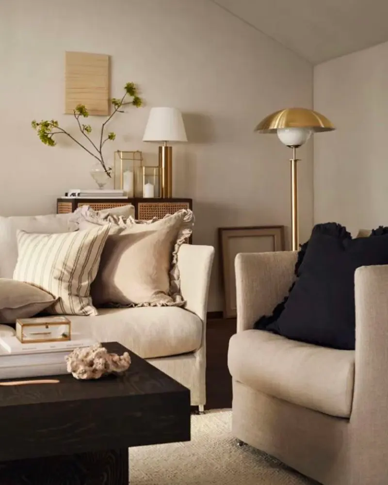 Chic beige living room with a brass floor lamp, cream armchairs, striped pillows, a black ruffled cushion and a dark coffee table