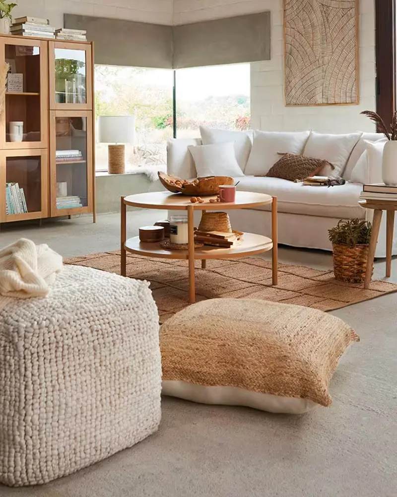 Cozy living room with a chunky knit pouf, a jute floor cushion, a white sofa, a round wood coffee table and a glass-front cabinet