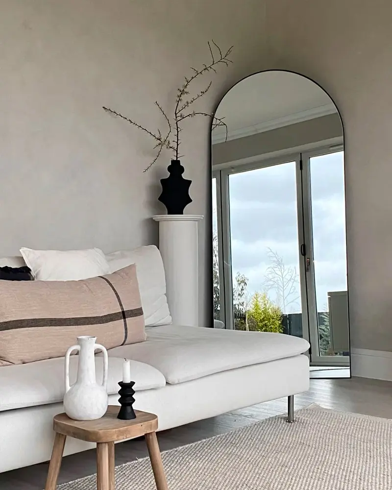 Minimalist beige living room with a white sofa, a striped lumbar pillow, a tall arched floor mirror and black and white vases