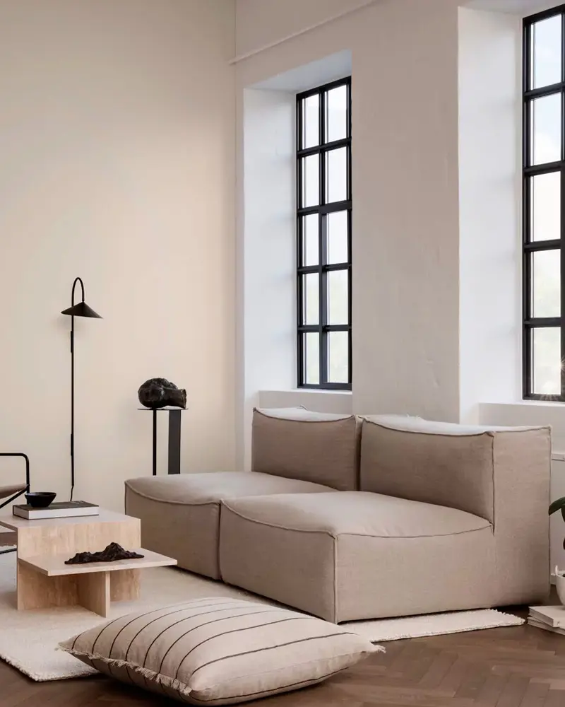 Minimal living room with a taupe modular sofa, a black floor lamp, floor cushions and tall black-framed windows