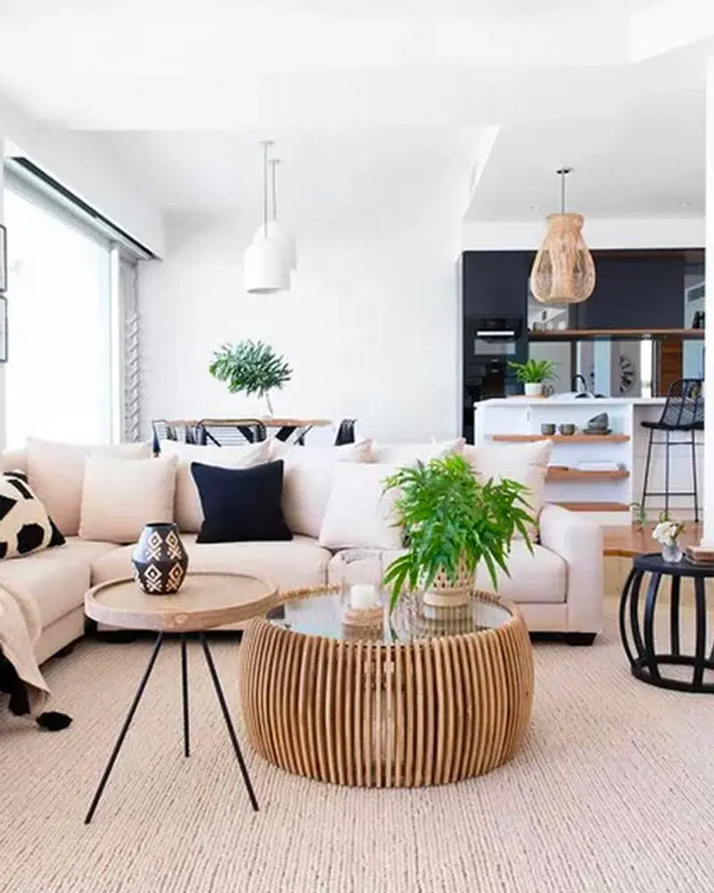 Open-plan living room with a cream sectional, a round wood slat coffee table, a glass-topped side table, rattan pendants and a fern