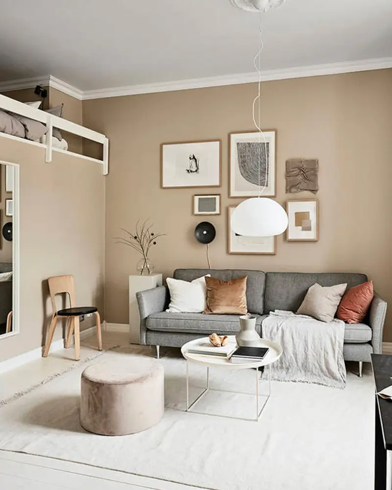 Living room with warm beige walls, a gallery of framed prints, a gray sofa with rust pillows, a round side table and a velvet pouf
