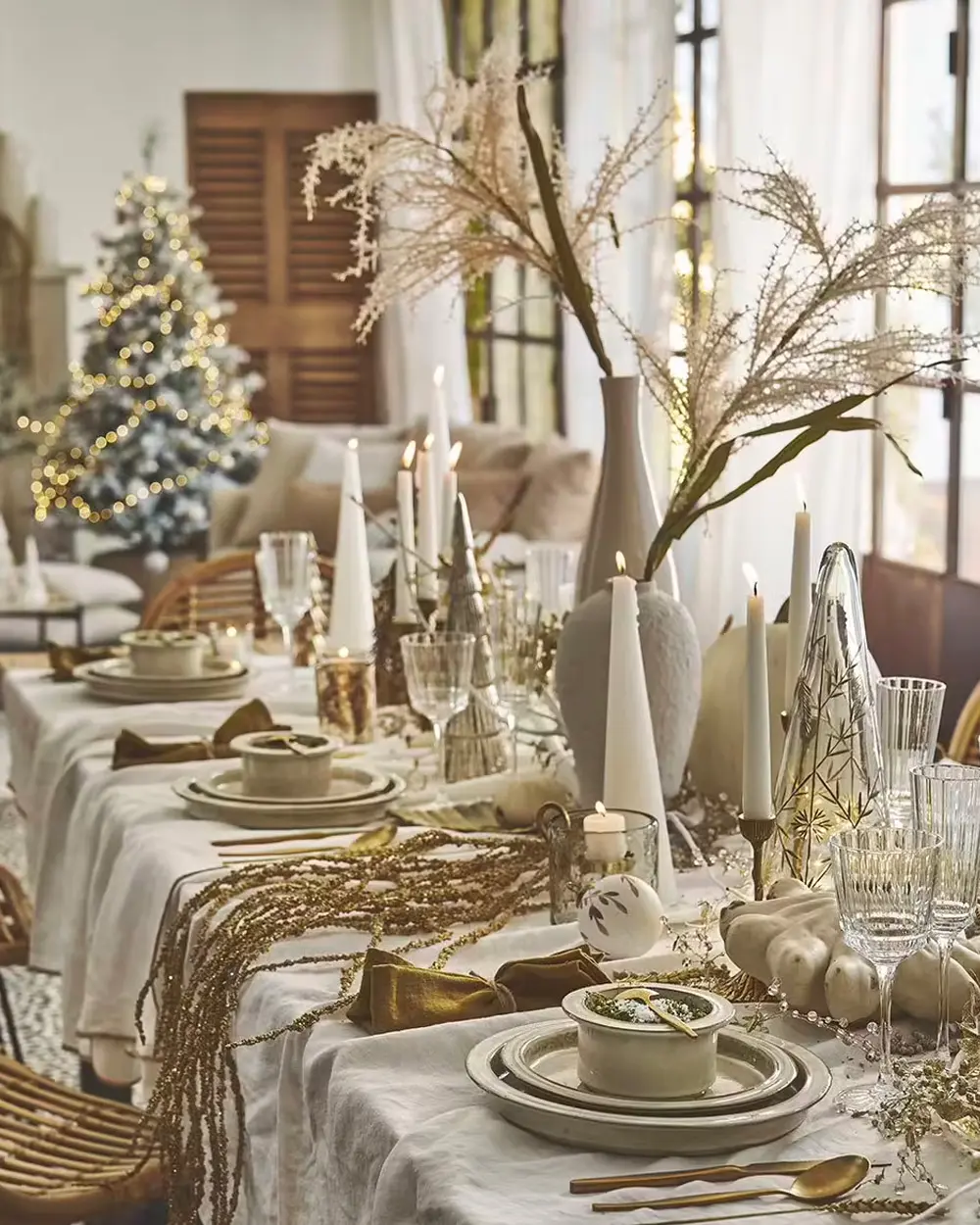 Beige Christmas table with tall pampas grass, white ceramic vases, candles and a lit tree behind