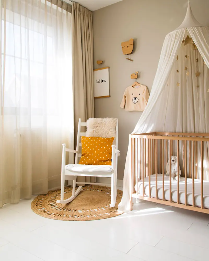 Beige nursery with canopy over a wooden crib, white rocking chair with mustard pillow and jute rug