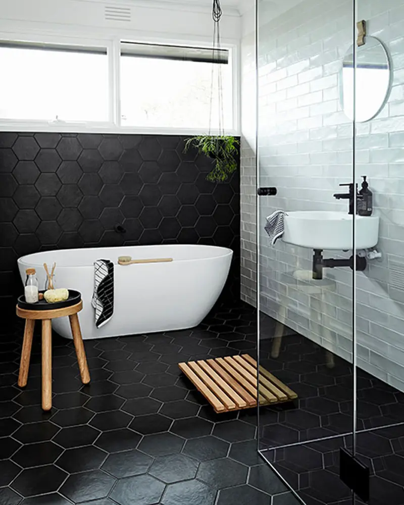 Freestanding tub on black hexagon floor with wood stool, slatted bath mat and glass shower