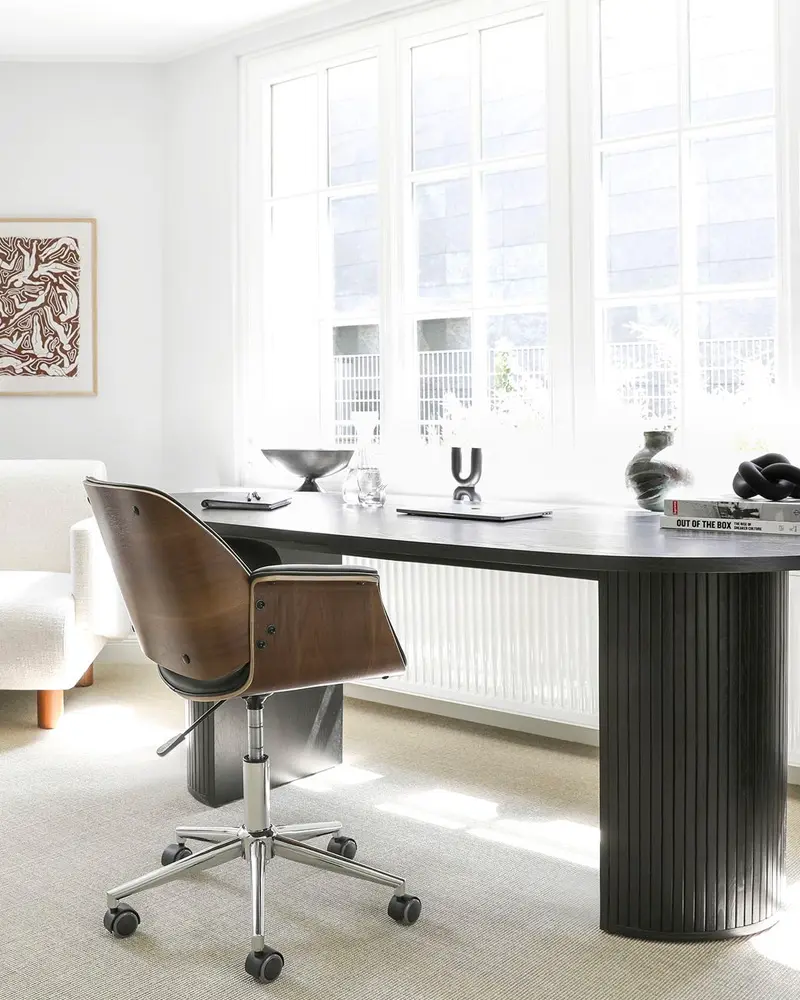Black fluted oval desk with walnut and chrome swivel chair by tall windows