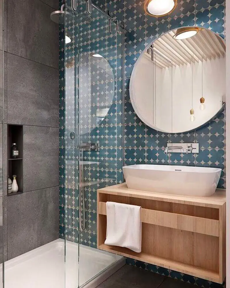 Floating wood vanity with white vessel sink, round mirror and blue patterned tiles behind a glass shower