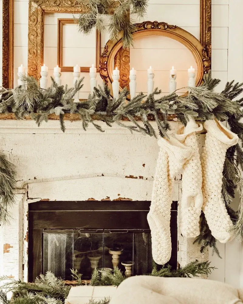 Chippy white mantel with layered gilded frames, a fir garland, taper candles and chunky knit stockings