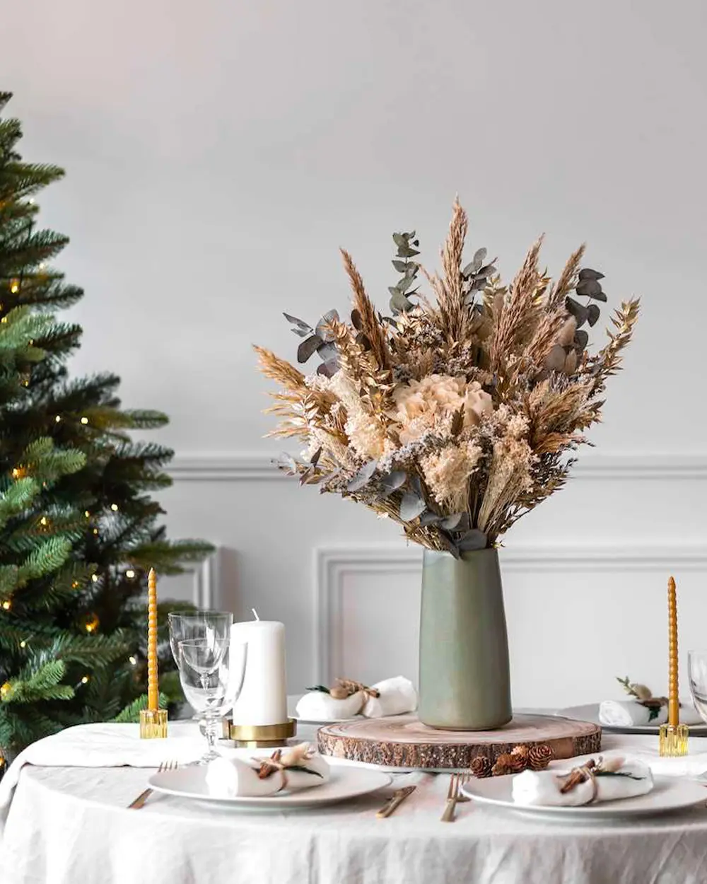 Round table with a big bouquet of dried grasses and eucalyptus in a green vase beside a tree