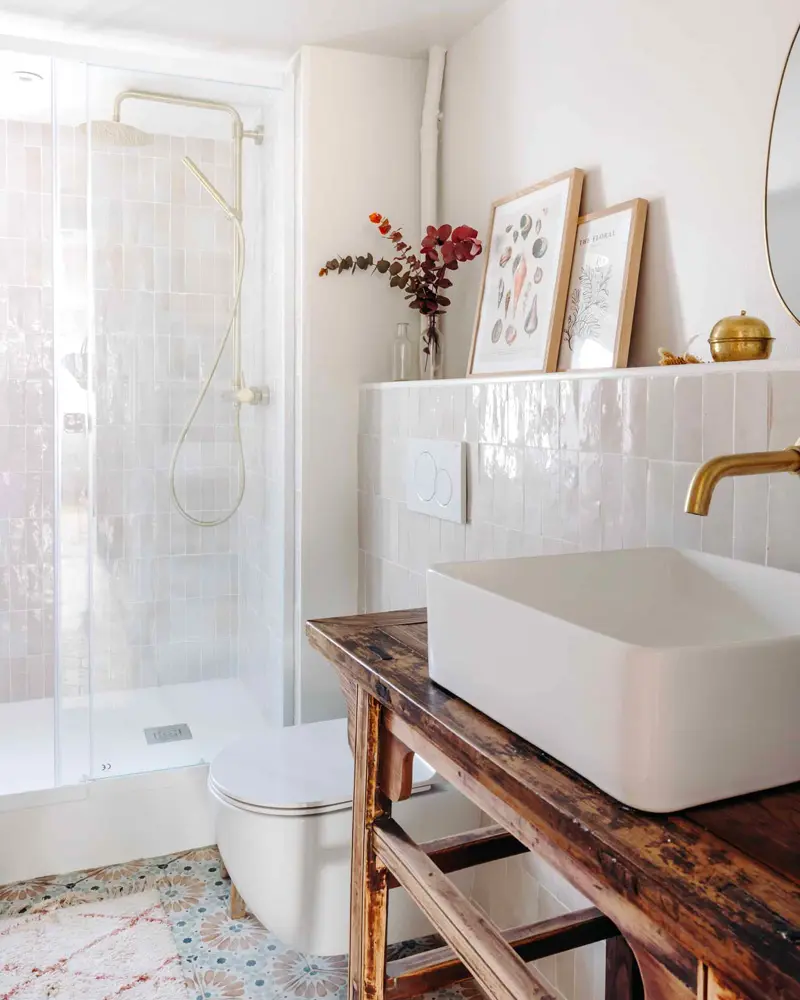 Square white vessel sink with a brass faucet on a rustic dark wood console, a glass shower and framed art on the ledge