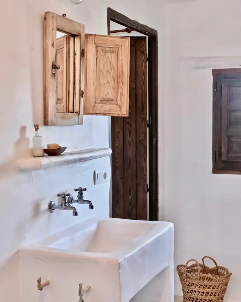 White limewashed bathroom with a built-in plaster sink, an old wooden wall cabinet with open doors and a dark wood door