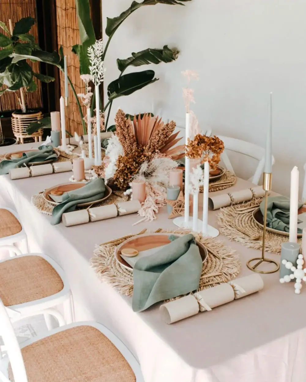 Long white table with woven placemats, sage napkins, crackers, dried palm and tall white candles