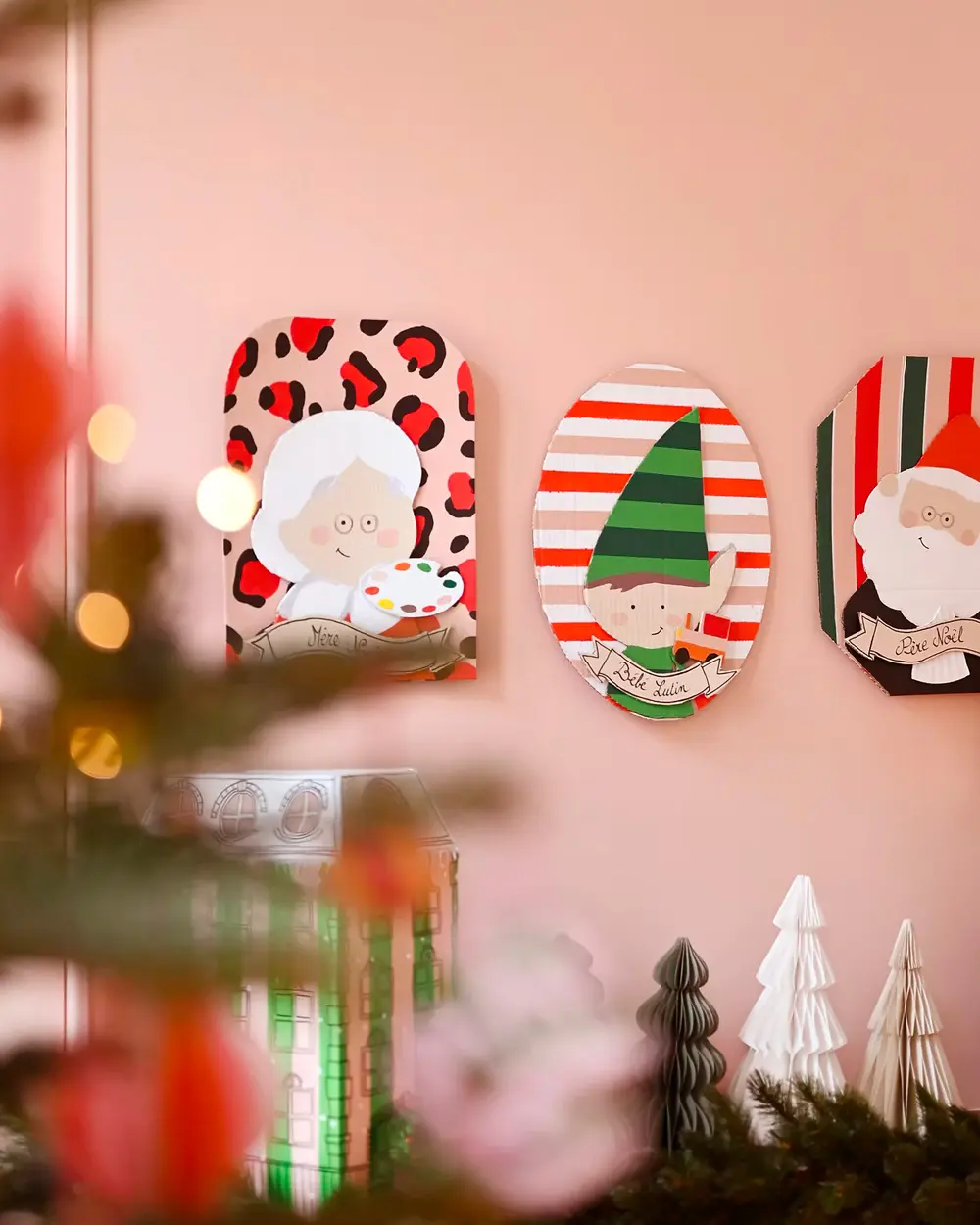 Cardboard portraits of Mrs. Claus, an elf and Santa on patterned backgrounds on a pink wall
