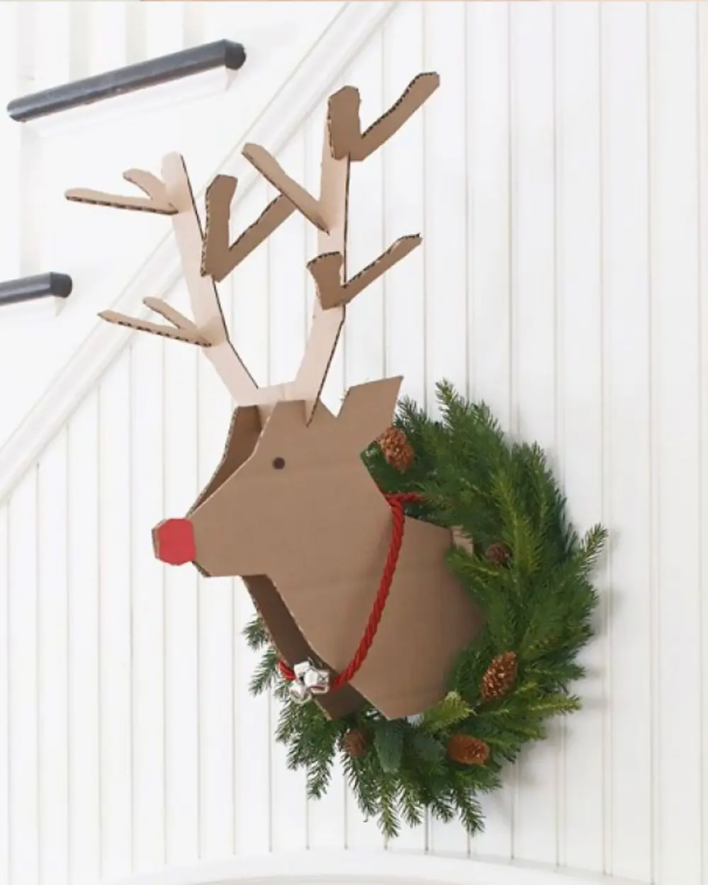 Cardboard reindeer head with antlers and a red nose wearing a fir wreath on a white wall