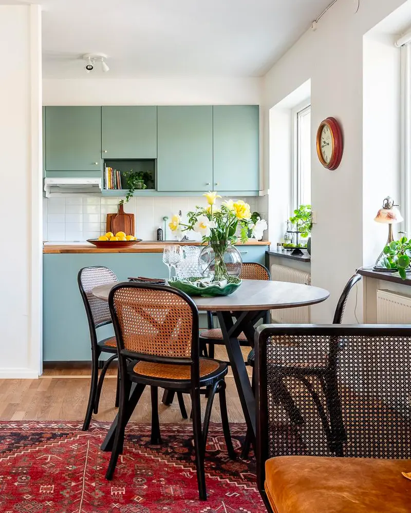 Round table with black cane chairs, red Persian rug, white flowers and sage kitchen cabinets