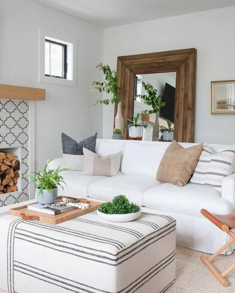 White coastal living room with a white sofa, a large reclaimed wood framed mirror, a striped upholstered ottoman, plants and a stacked firewood niche