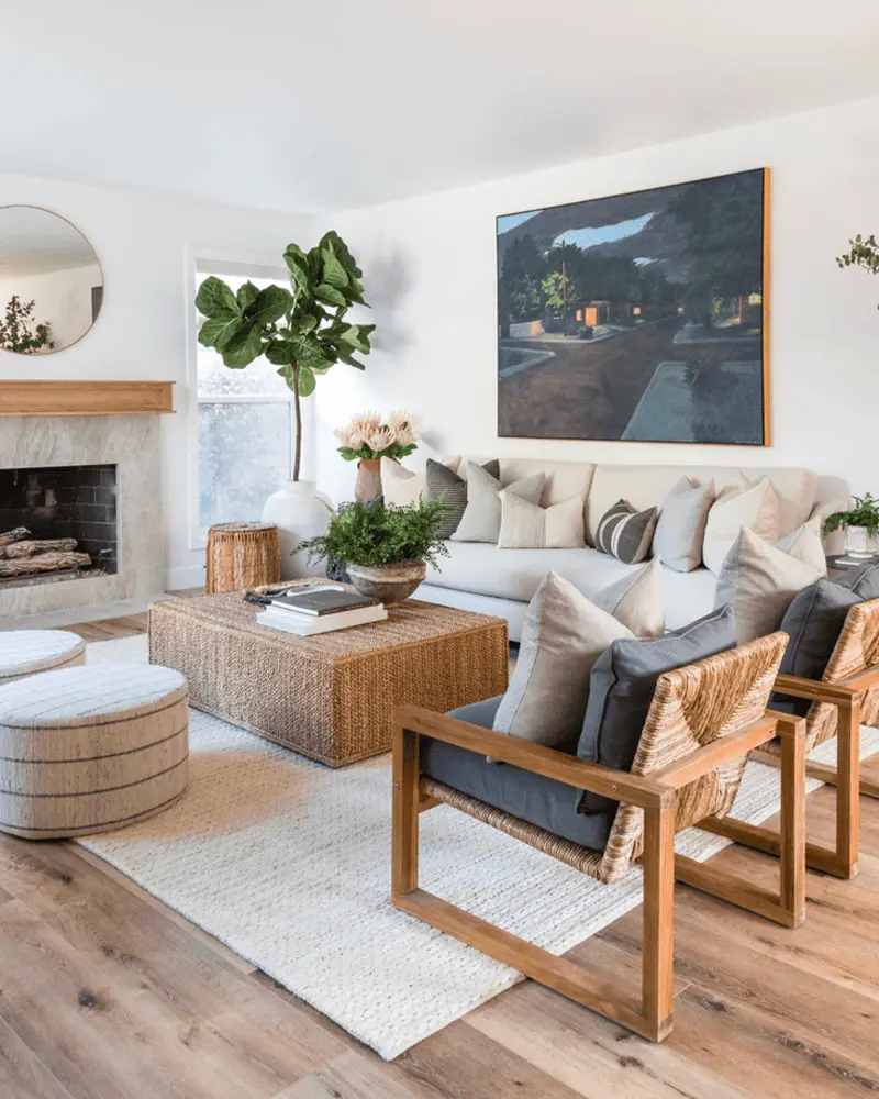 Coastal living room with a beige sofa, a woven coffee table, wood armchairs with navy cushions, a fiddle leaf fig, a fireplace and a dark landscape painting