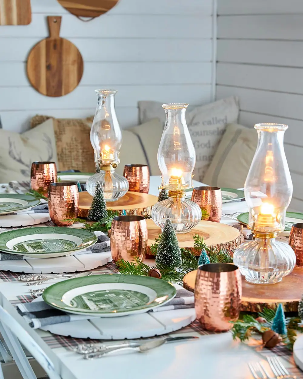 Table with glass oil lamps on wood rounds, copper cups, green transferware plates and bottle-brush trees