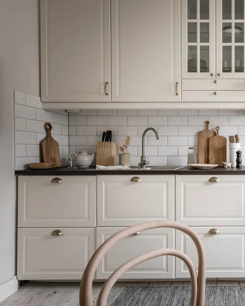 Greige shaker cabinets with glass-front upper, brass cup pulls, subway tile and bentwood chair