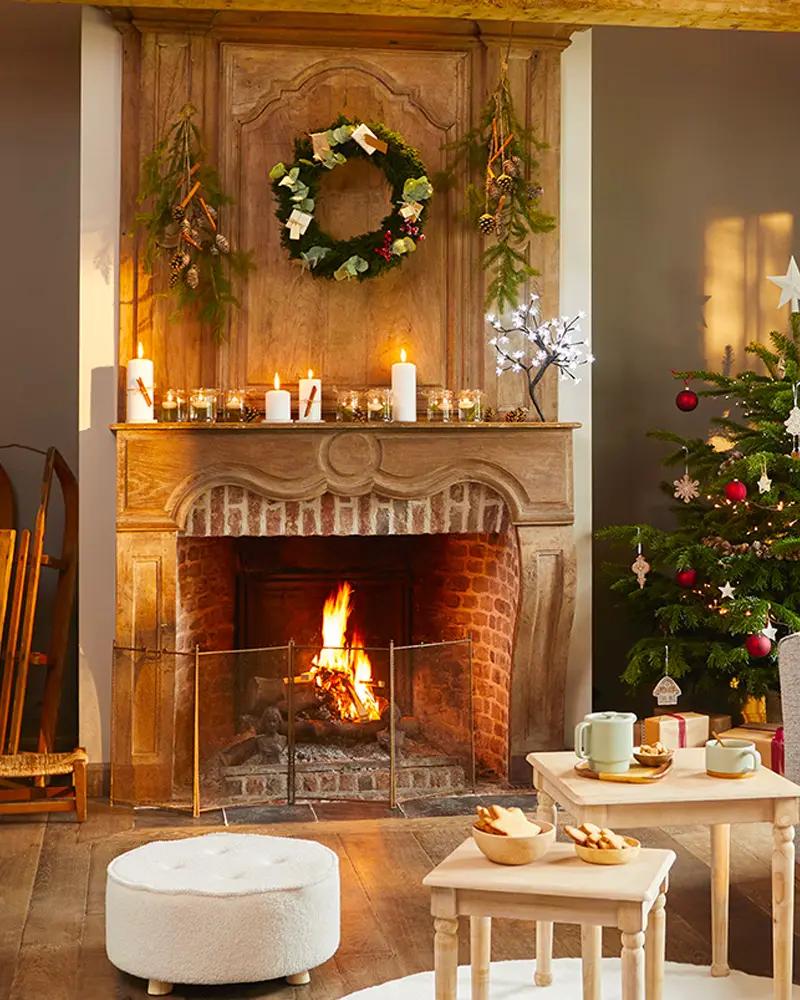 Carved wood fireplace with a wreath, hanging greenery and candles, a lit fire and a decorated tree