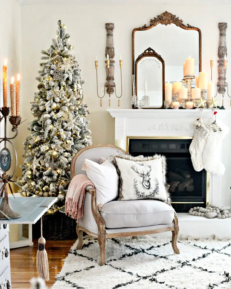White mantel with a gilded mirror, tall candles and a flocked tree beside a carved armchair
