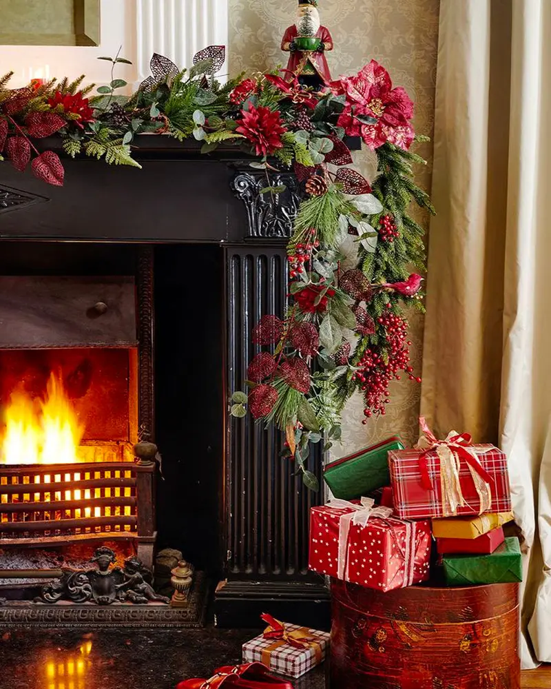 Black fireplace draped with a lush red and green garland, a roaring fire and wrapped gifts