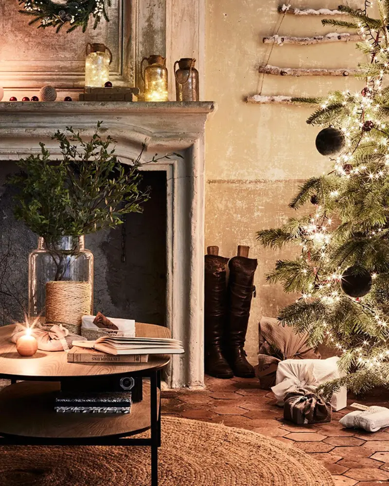 Aged stone fireplace with greenery in a glass vase, jars, a lit tree and boots by a round coffee table