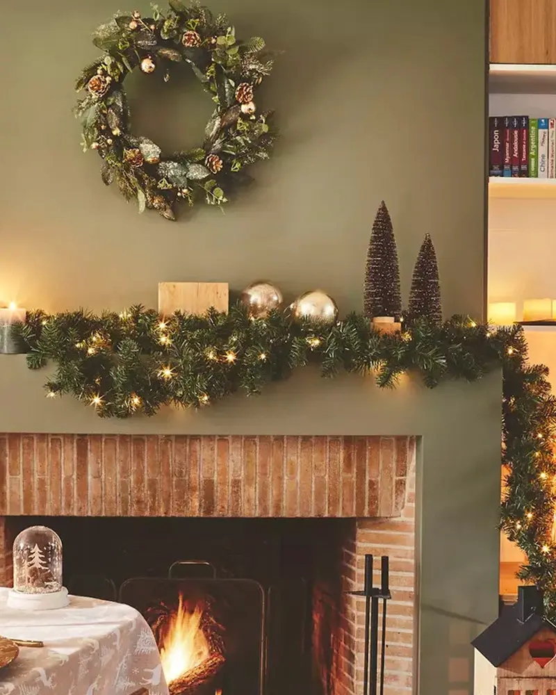 Brick fireplace on a sage green wall with a wreath and a lit garland topped with gold ornaments