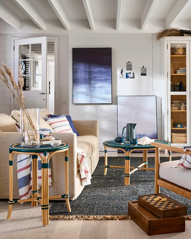 Coastal living room with a beige sofa, striped pillows, rattan bistro side tables, a blue abstract painting and a dark rug