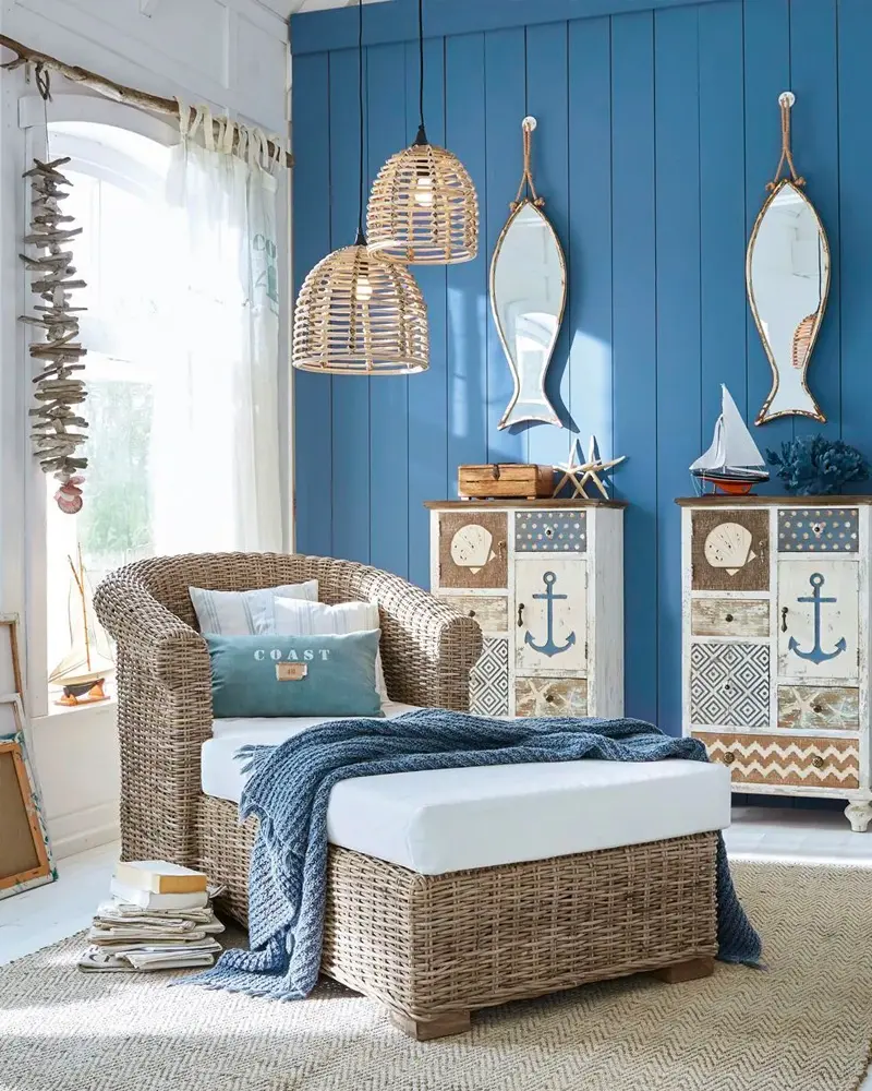 Blue paneled wall with fish-shaped mirrors, wicker pendants, a wicker armchair and ottoman and white cabinets with anchor motifs