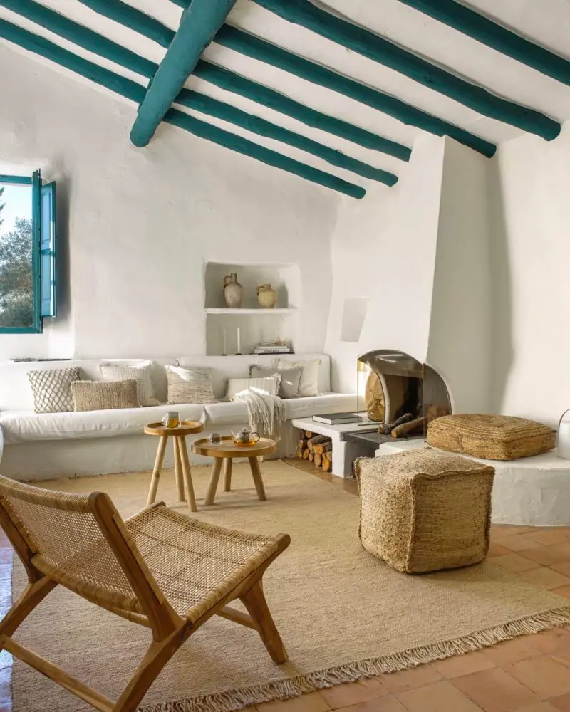 Whitewashed living room with a built-in plaster sofa, teal-painted ceiling beams, a woven lounge chair, jute poufs and a fringed rug