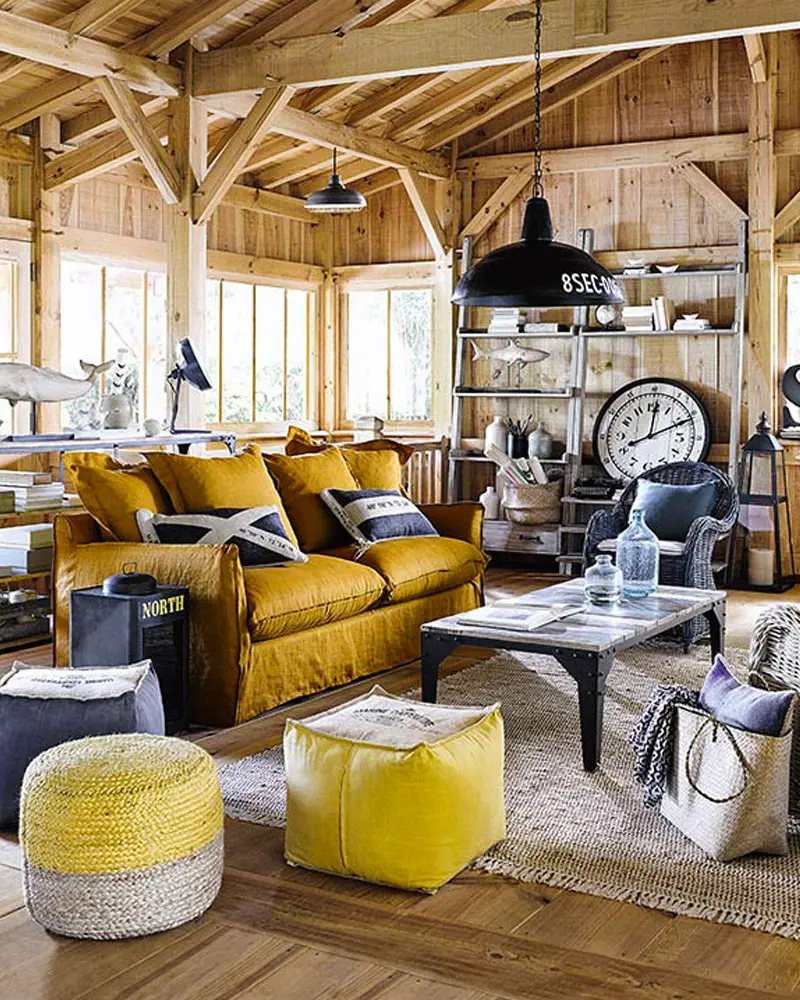 Wood-paneled cabin living room with a mustard velvet sofa, yellow knit poufs, a black industrial pendant and a large wall clock