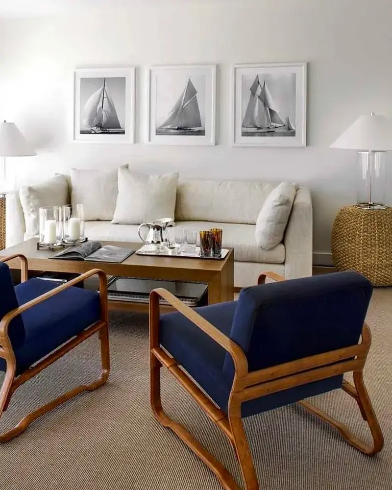 Chic coastal living room with a cream sofa, two navy armchairs with wood frames, a sisal rug and three framed black and white sailboat prints