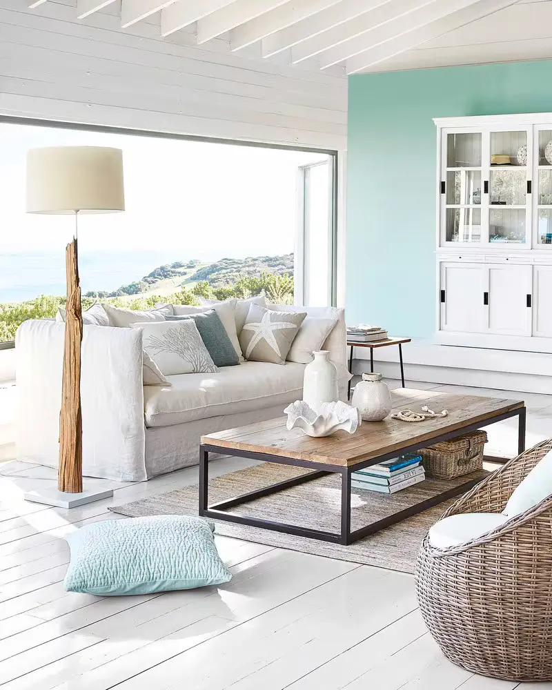 Living room with an ocean view, a white sofa with blue pillows, a driftwood floor lamp, a wood and metal coffee table, a seafoam wall and a shell bowl