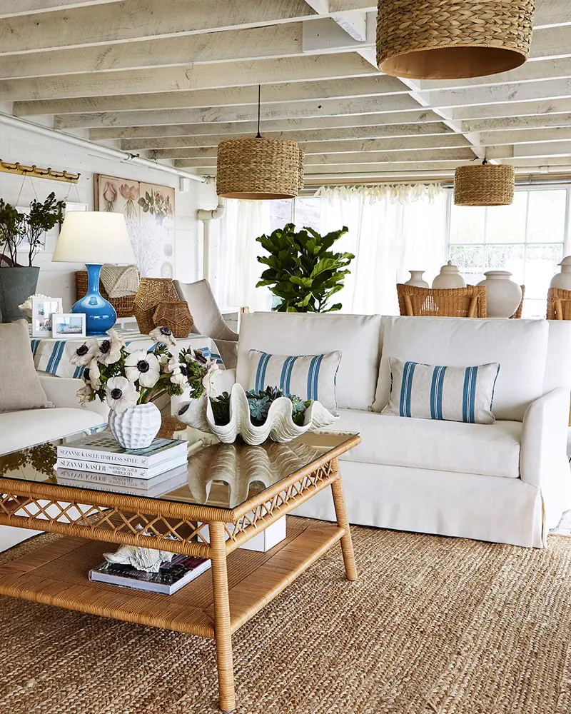 Coastal living room with a white slipcovered sofa, blue striped pillows, a two-tier rattan coffee table, a giant clam shell and woven pendants
