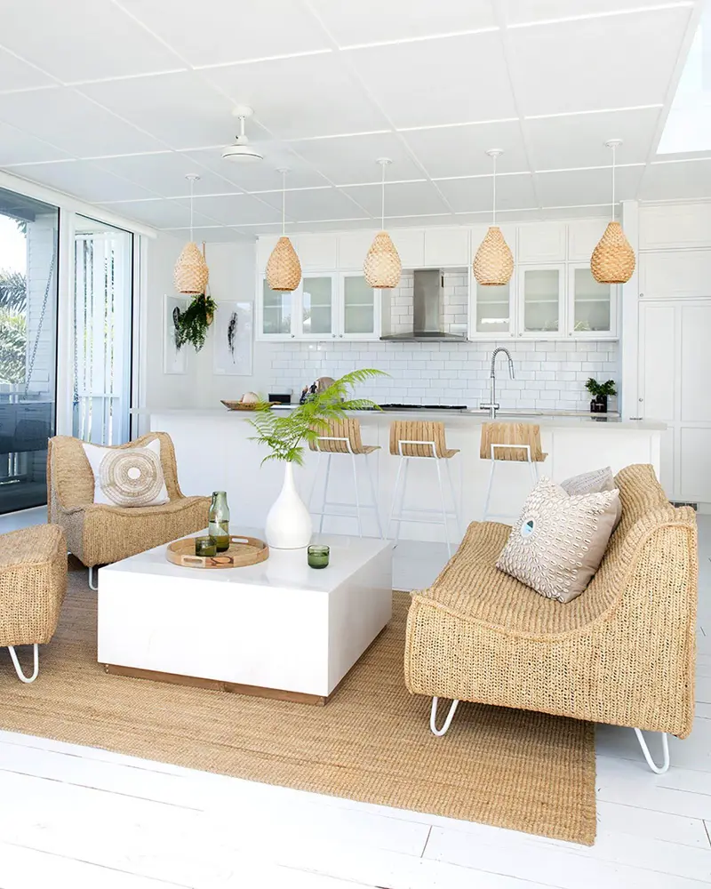Bright white coastal living room with a woven rattan sofa and armchairs on hairpin legs, a white cube coffee table, a jute rug and rattan pendants