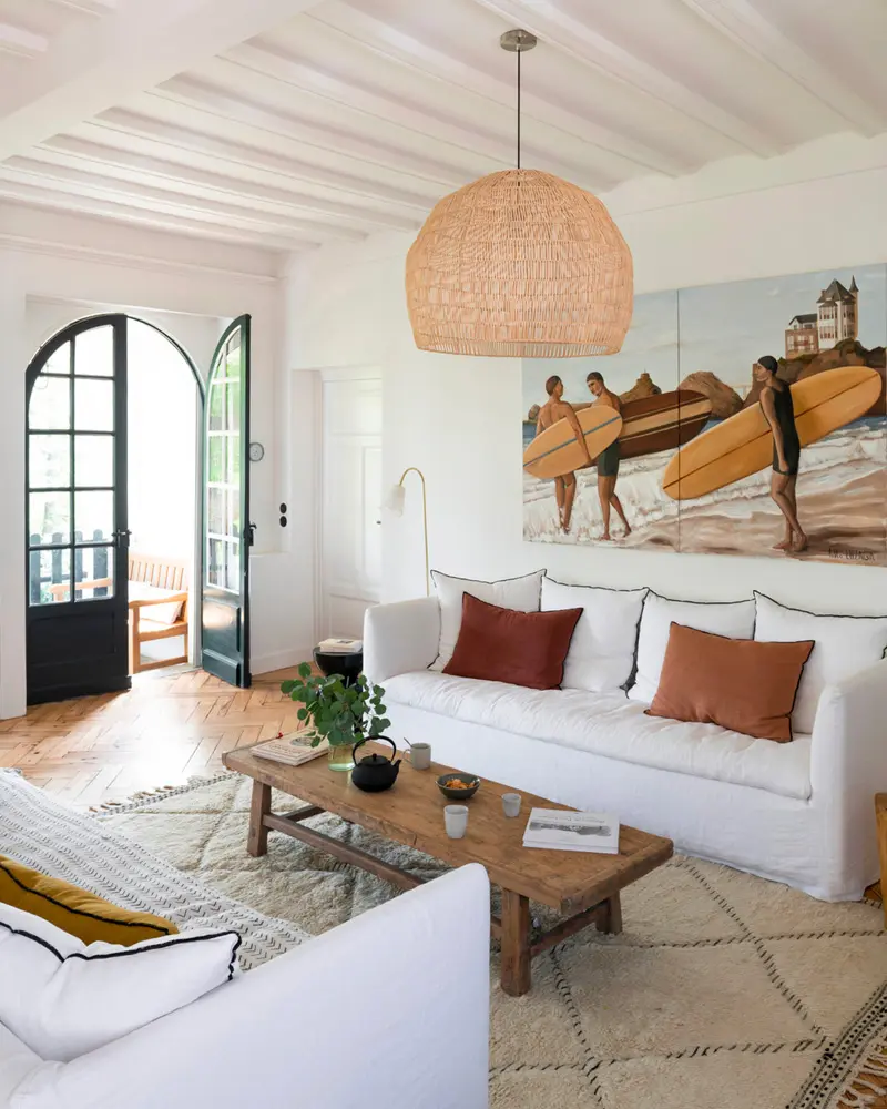 White coastal living room with slipcovered sofas, rust pillows, a large surfing painting, a woven dome pendant and an arched glass door