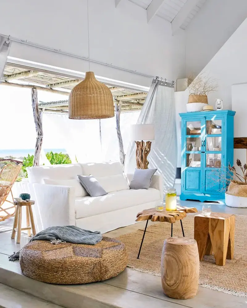 Living room open to the ocean with a white sofa, a rattan pendant, a turquoise painted cabinet, tree-stump side tables and a round jute pouf