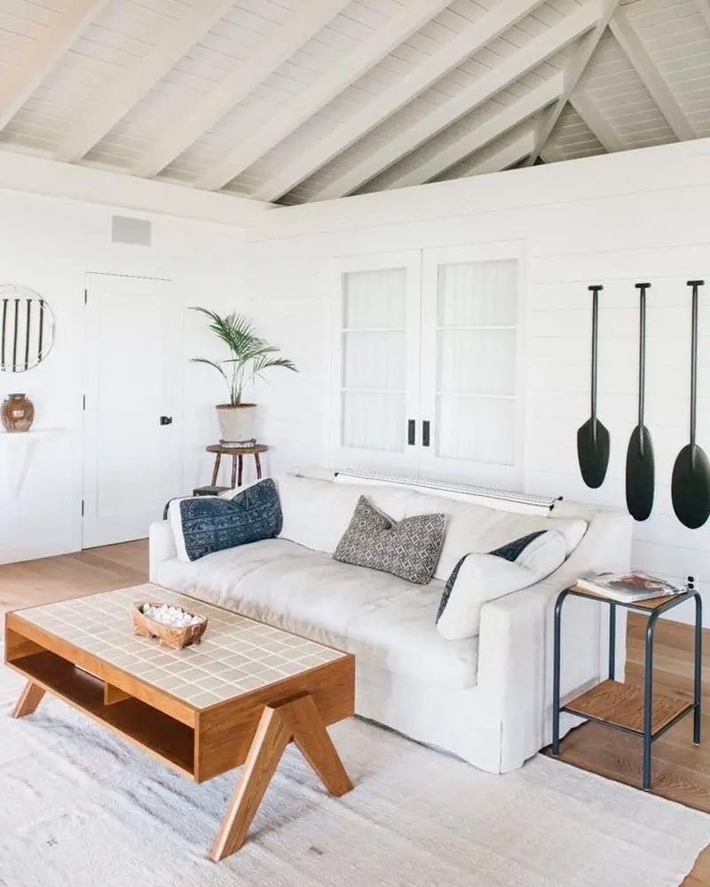 All-white coastal living room with a vaulted white ceiling, a white sofa, a mid-century wood coffee table and three black oars on the wall