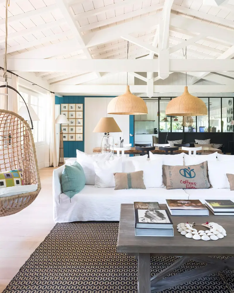 Open coastal living room with a white slipcovered sofa, woven rattan pendants, a hanging egg chair, a geometric rug and a white beamed ceiling