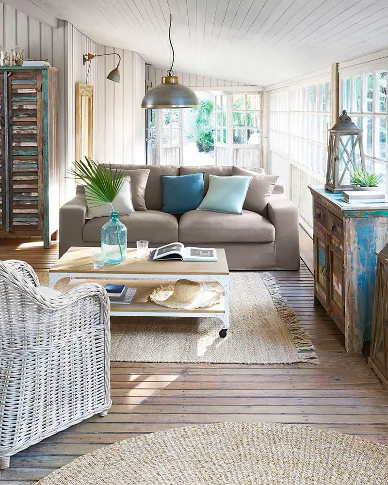 Coastal living room with weathered wood floors, a taupe sofa with turquoise pillows, a white wicker armchair, a jute rug and a metal pendant