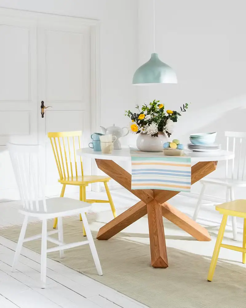 Round wood table with crossed legs, white and yellow chairs, striped runner and mint pendant