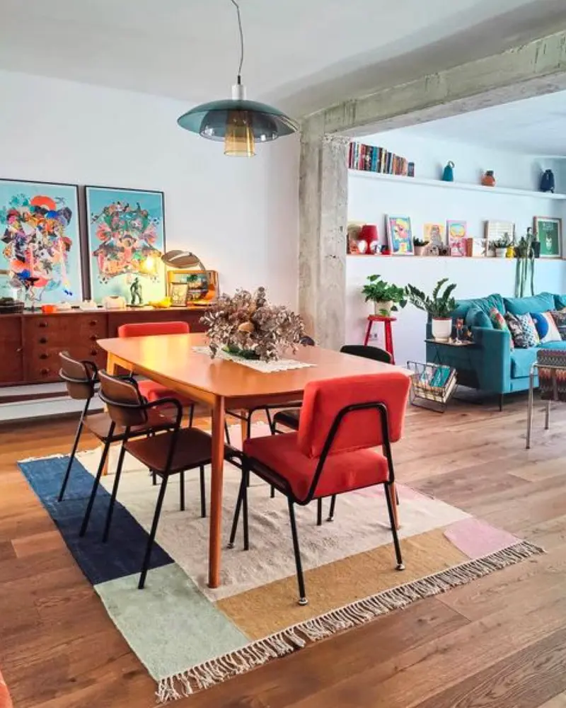 Teak dining table with a red armchair, orange chairs, a colorful rug and bright artwork