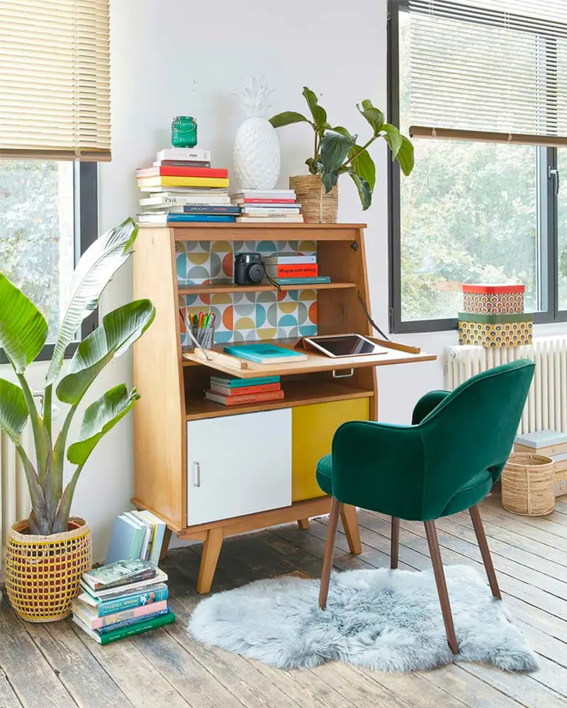 Vintage secretary desk with patterned interior, green velvet armchair, sheepskin and bird of paradise plant