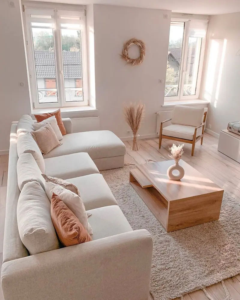 Cozy beige living room with a deep cream sectional, blush pillows, a wood coffee table, a dried flower wreath on the wall and a fluffy rug