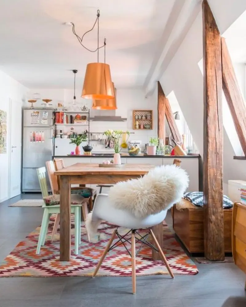Wood table with sheepskin on a white molded chair, orange pendant lights and colorful kilim rug under exposed beams