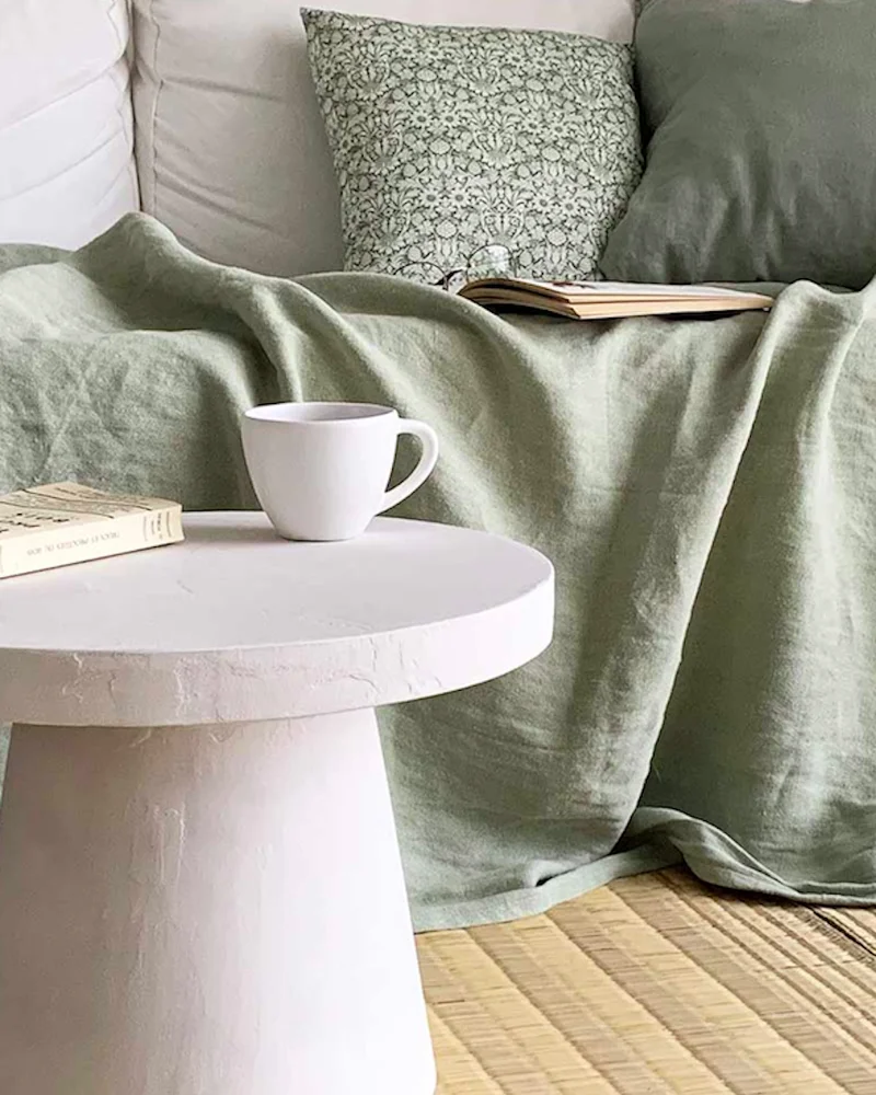 Round white concrete pedestal table holding a mug beside a green linen throw